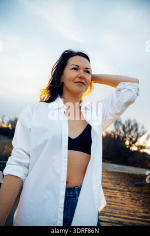 Frau mit einem ruhigen Ausdruck steht bei Sonnenuntergang draußen, trägt ein weißes Hemd und legere Kleidung. Das warme Leuchten des Sonnenuntergangs unterstreicht ihre Rela Stockfoto