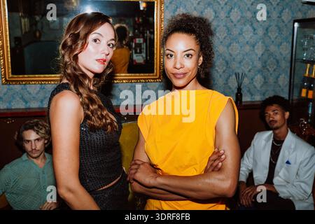 Gruppe von verschiedenen Freunden, die sich in einer stilvollen Bar treffen und feiern, zwei Frauen stehen im Vordergrund mit Männern, die sich hinter ihnen entspannen, en Stockfoto