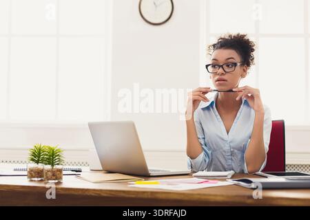 Frau sitzt am Schreibtisch mit Laptop und Schreibwaren, während sie in einem Büro nachdenkt Stockfoto