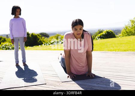 Mutter übt Yoga an Deck, während Tochter zusieht und sich im Freien entspannt Stockfoto