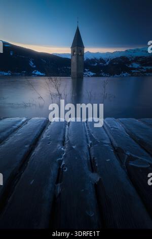 Der Blick auf einen einsamen Kirchturm erhebt sich aus dem eisigen Griff eines gefrorenen Sees, eingerahmt von schneebedeckten Bergen unter einem Dämmerungshimmel, Reschen, Trentino-Süd-Tyro Stockfoto