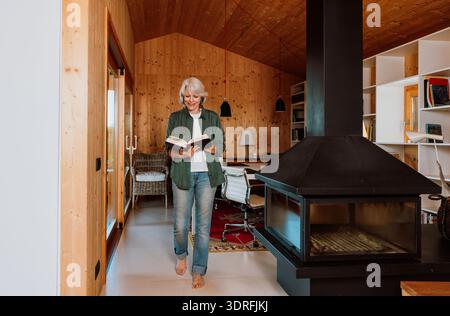 Eine Seniorin steht barfuß, lässig gekleidet, liest ein offenes Buch in einem komfortablen Wohnbereich mit modernem Kamin und Holzwänden, genießt es Stockfoto