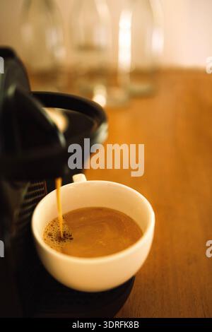 Nahaufnahme einer weißen Tasse gefüllt mit köstlichem aromatischem Kaffee mit Milch. Die Kaffeemaschine steht auf einem Holztisch und macht einen Stockfoto