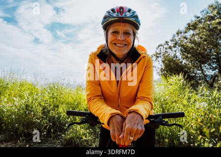 Ältere Frau mit Helm und gelber Jacke, die sich während einer sonnigen Outdoor-Aktivität glücklich auf ihrem Fahrradlenker ausruht und einen aktiven und ihn umschließt Stockfoto