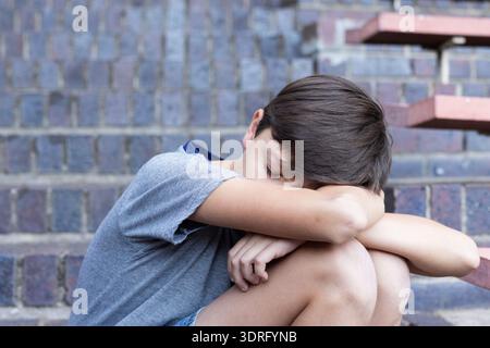 Auf Stufen sitzend, mit dem Kopf auf den Armen, traurig in der Schule. Emotional, allein, Kontemplation, Kindheit, Stress, Einsamkeit Stockfoto