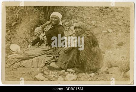 Diese Schwarzweiß-Postkarte aus dem Jahr 1913 zeigt zwei Pala-Frauen (Mission Indian), die draußen auf einem felsigen Hügel im San Diego County, Kalifornien, sitzen. Die Frauen tragen traditionelle Kleidung und das Bild hebt die Fotografie des frühen 20. Jahrhunderts hervor, indigene Darstellung, kulturellen Kontext, Landschaft, soziale Umgebung, und ethnographische Dokumentation der Pala. Stockfoto