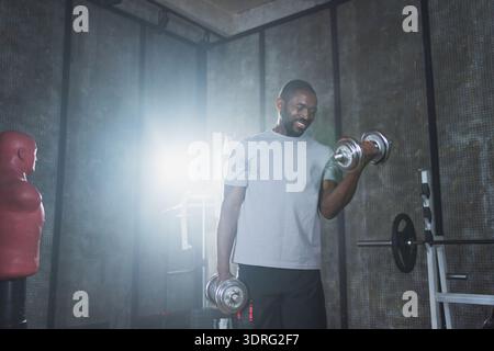 Fitness-Workout im Fitnessstudio. Afroamerikaner Bodybuilder, der im Fitnessstudio Kurzhanteln aufnimmt. Workout Gewichtstraining Bizepsmuskeln mit Stockfoto