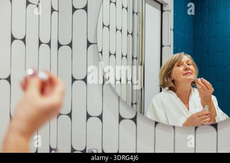 Eine ältere Frau in einem weißen Frottee-Morgenmantel trägt ein Serum auf ihr Gesicht. Eine reife Dame kümmert sich um ihre Haut und schaut h an Stockfoto