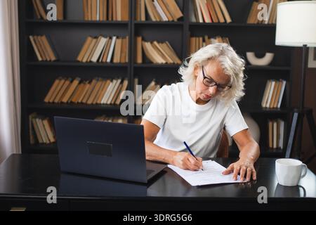 Ältere Frau mittleren Alters, die mit Laptop am Arbeitsplatz Notizen schreibt. Konzentrierte, reife Geschäftsfrau arbeitet im Büro Stockfoto