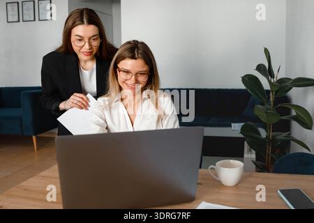 Zwei Frauen arbeiten an einem Computer in einem Büro zusammen, während sie an einem Schreibtisch mit einer Kaffeetasse in der Nähe über Arbeitsaufgaben diskutieren. Stockfoto
