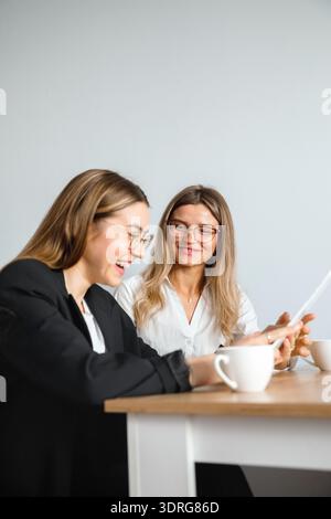 Zwei Frauen sitzen an einem Tisch in einem Büro und diskutieren über Arbeit, während sie sich ein Dokument ansehen und Kaffee in Tassen trinken. Stockfoto