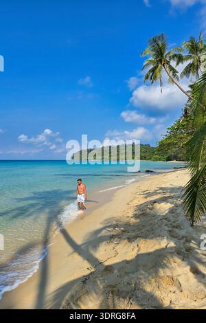 Ein Alleinreisender spaziert entlang eines ruhigen Sandstrandes, während ihm sanfte Wellen zu Füßen liegen. Üppige Palmen wiegen in der sanften Brise und schaffen ein friedliches atmos Stockfoto