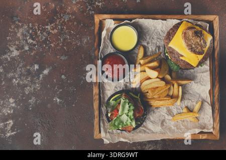 Leckerer Burger mit Käse, Pommes und Saucen, serviert auf einem Holztablett Stockfoto