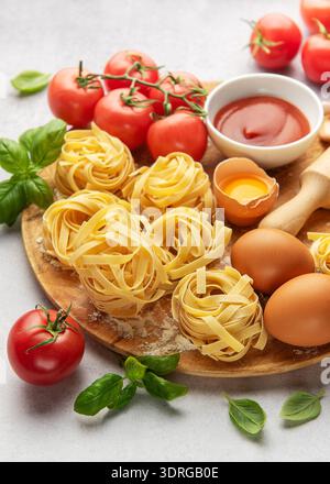 Frische Tagliatelle-Pasta nestet mit Tomaten, Basilikum, Eiern, Mehl und Tomatensauce auf Holzbrett Stockfoto
