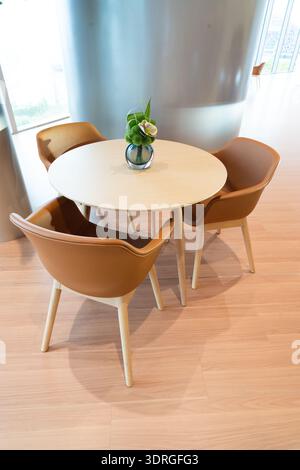 Moderner runder Holztisch mit vier braunen Ledersesseln in minimalistischem Innenraum, hellem Holzfußboden und großer Fensterkulisse Stockfoto