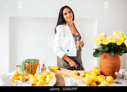 Eine junge schwangere Frau lächelt und hält ihren Bauch in der Küche. Lächelnde werdende Mutter steht am Tisch mit Zitronen im Weinglas Stockfoto
