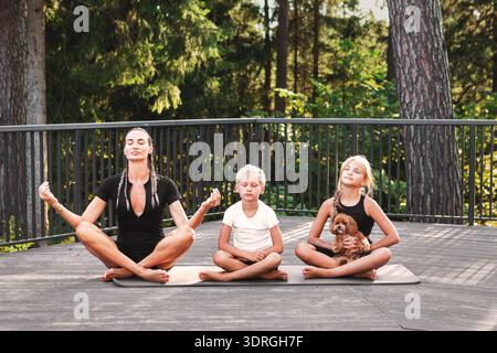 Achtsame Mutter übt Meditation mit Kindern und einem kleinen Hund auf einer Holzterrasse im Freien. Ruhiges Familienerlebnis, Yoga und Achtsamkeit Stockfoto