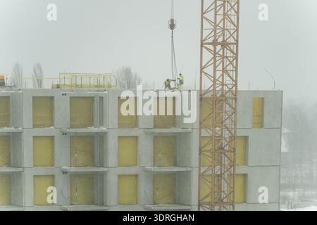 Modernes Wohnprojekt mit Betonfertigfassade, gestapelten Balkonen, Kranturm und Dacharbeitern, die Bauelemente installieren Stockfoto