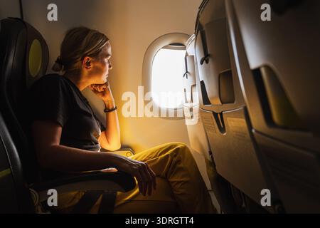 Glückliche Passagierin fliegt im Flugzeug. Mädchen, das im Flugzeug sitzt und aus dem Fenster blickt und auf Urlaubsreise geht. Unterwegs f Stockfoto