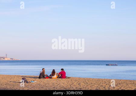 Malaga, Spanien – 11. Februar 2020: Eine Gruppe von Freunden sitzt am Sandstrand von Malagueta unter klarem blauen Himmel und genießt die malerische Aussicht auf den ruhigen Medit Stockfoto