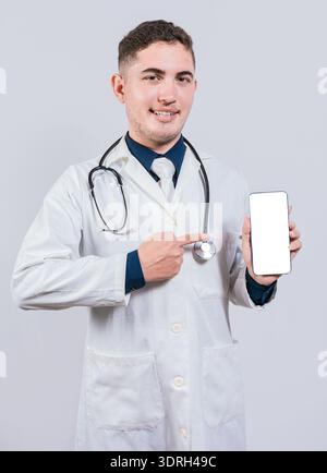 Fröhlicher Arzt, der auf den Bildschirm des Mobiltelefons zeigt. Junger Arzt zeigt und zeigt isoliert auf den Smatphone-Bildschirm Stockfoto