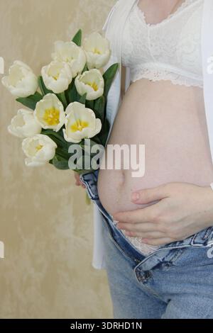Eine Frau zeigt ihr Baby, während sie einen Strauß weißer Tulpen hält. Sie trägt lässigen Denim und ein leichtes Oberteil, was für einen fröhlichen Moment sorgt. Stockfoto