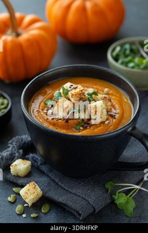 Scharfe Kürbiscremesuppe in schwarzem Becher, serviert mit Kürbiskernen und Croutons, dunkler Hintergrund Stockfoto