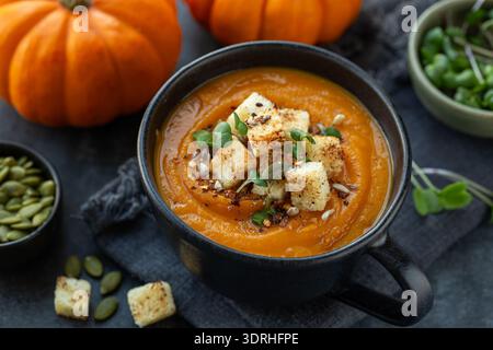Scharfe Kürbiscremesuppe in schwarzem Becher, serviert mit Kürbiskernen und Croutons, dunkler Hintergrund Stockfoto