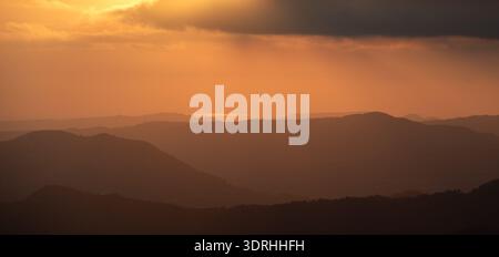 Schichten ferne Bergsilhouetten unter einem warmen orangen Himmel bei Sonnenuntergang schaffen eine ruhige und weite natürliche Landschaft. Stürmischer, dramatischer Himmel Stockfoto