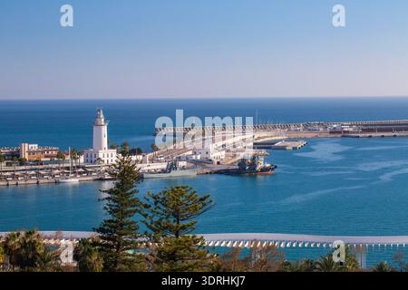 Malaga, Spanien. Der weiße Leuchtturm La Farola am Eingang zum Hafen von Malaga mit angedockten Schiffen und Industriekränen. Mediterrane Küstenlandschaft und Stockfoto