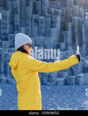 Ein Reisender in einer leuchtend gelben Jacke macht ein Selfie am Strand von Reynisfjara, umgeben von einzigartigen Basaltsäulen. Das ist atemberaubend Stockfoto