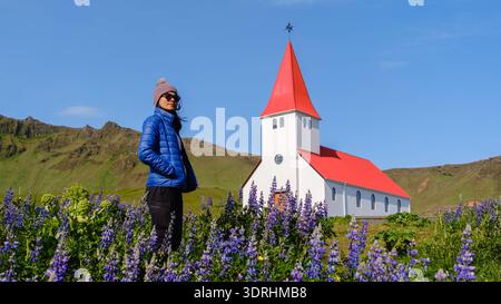 Eine asiatische Reisende steht inmitten eines blühenden Lupinenfeldes in der Nähe der bezaubernden Kirche Vik in Island. Stockfoto