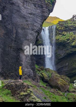 Eine Frau in einer leuchtend gelben Regenjacke bildet einen wunderschönen Kontrast zu den felsigen Klippen und dem kaskadierenden Kvernufoss-Wasserfall. Frau in Stockfoto