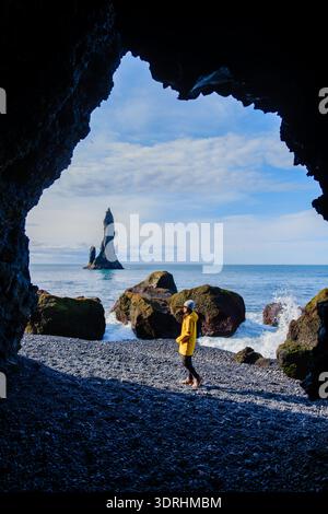 Eine Frau, die in einer felsigen Höhle steht, bewundert den atemberaubenden Strand von Reynisfjara. Die dramatischen Seestapel steigen majestätisch auf Stockfoto