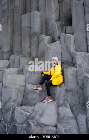 Eine Frau in gelber Regenjacke und fesselnden Granitsäulen erhebt sich vom Strand Reynisfjara mit einem Reisenden in leuchtend gelber Kleidung Stockfoto