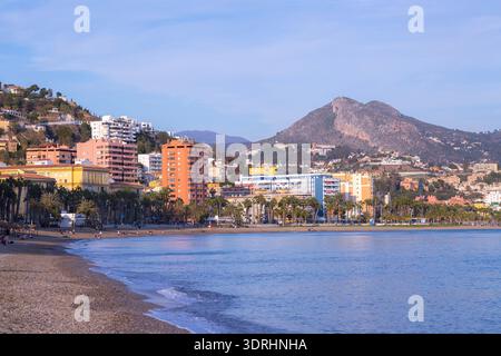 Malaga Spanien Weitwinkelblick auf den Strand La Malagueta bei Sonnenuntergang mit blauen Meereswellen, die auf dem Kieselstrand Rollen, und Stadtgebäuden unter einem klaren Himmel in t Stockfoto