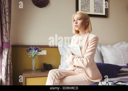 Frau in einem rosafarbenen Anzug sitzt tagsüber auf einem Bett mit einem Tablet in einem Hotelzimmer Stockfoto