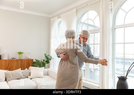 Bleiben Sie in Bewegung. Ein romantisches älteres Ehepaar, das zu Hause zu Musik tanzt. Glücklich lächelnde Familie Rentner Frau Ehemann Ehefrau Stockfoto