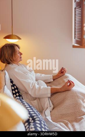 Eine ältere Frau im weißen Mantel sitzt im Bett und meditiert Stockfoto