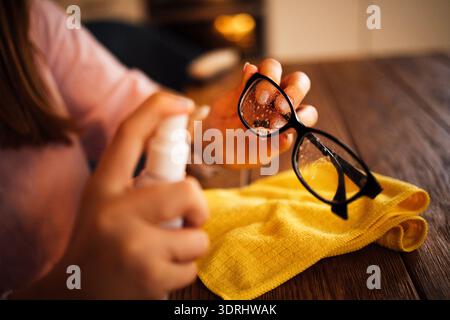 Eine Frau sprüht Reinigungslösung auf die Brille. Weibliche Hände halten eine Brille und eine Flasche mit Reinigungsspray. Ein gelbes Tuch und ein Stockfoto