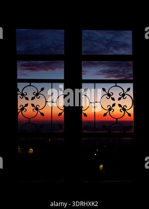 Blick auf den Sonnenuntergang durch kunstvoll verzierte Fenster in der Toskana, Italien, dramatischer orange-blauer Himmel, eingerahmt von dekorativer Silhouette aus Eisen über sanften Hügeln. Stockfoto