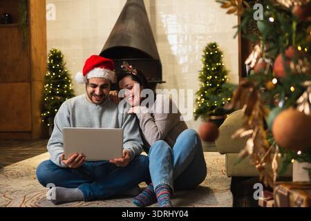 Frohe Weihnachten glückliches junges erwachsenes Paar mit Laptop in der Nähe von Weihnachtsbaum und Kamin. Glückliche Familienfrau, die auf der Suche nach dem Kauf ist Stockfoto