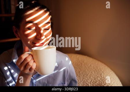 Ein charmantes Mädchen in einem blauen Hemd sitzt in einem Sessel und genießt einen köstlichen Kaffee. Ein Student trinkt Tee und lächelt Ho an Stockfoto