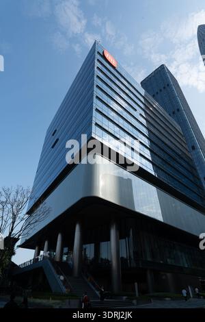 Moderner Glashochhaus mit geschwungenem Sockel und orangefarbenem Logo unter blauem Himmel im urbanen Stadtbild Stockfoto