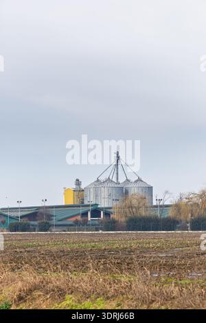 IVANKOVO, KROATIEN, 02.14.2026: Kornsilos und ein Metalllager erheben sich hinter einem ausgeernteten Feld unter einem hellen bedeckten Himmel und heben die Moderne hervor Stockfoto
