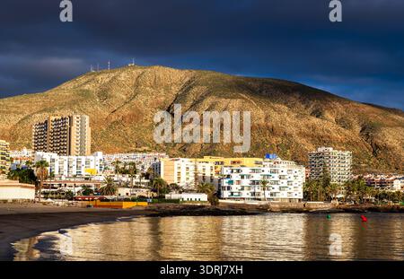 Blick auf den Strand Playa de Los Cristianos und den Berg Guaza auf Teneriffa, Kanarischen Inseln, Spanien. Mit Sandstrand, ruhigem Meereswasser und bei Sonnenuntergang beleuchteten Resortgebäuden. Stockfoto