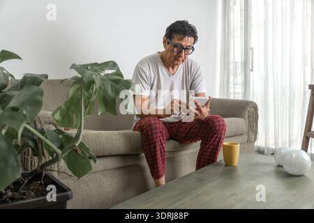 Mann, der sich auf dem Sofa mit Schlafanzug entspannt und zu Hause ein Tablet benutzt Stockfoto