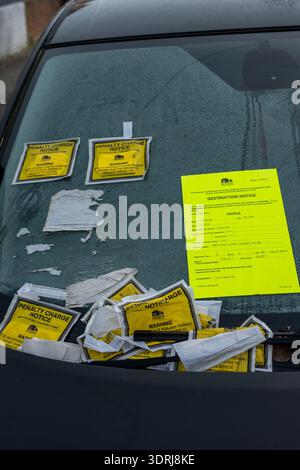 Auto mit mehreren Parktickets und festem Strafzettel neben einem Schild, das an der Windschutzscheibe befestigt ist und auf dem die Entfernung und Zerstörung des Fahrzeugs hingewiesen wird. Stockfoto