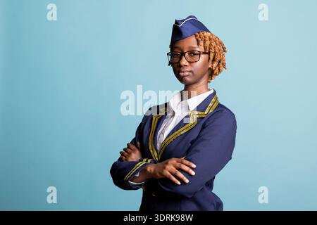 Porträt der Schwarzen-Luft-Gastgeberin in blau-goldenen Uniformen, die sich selbstbewusst kreuzen. Die junge Frau in Brille steht vor isoliertem Studiohintergrund und repräsentiert professionelle Fluggesellschaften. Stockfoto