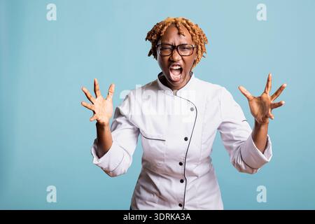 Frustrierte Küchenchefin in Uniform, die vor Wut und Wut vor blauem Hintergrund schreien. Junge professionelle Köchin, die schreien und extreme Frustration über schreckliche Mahlzeiten und Küchenstress freisetzen. Stockfoto
